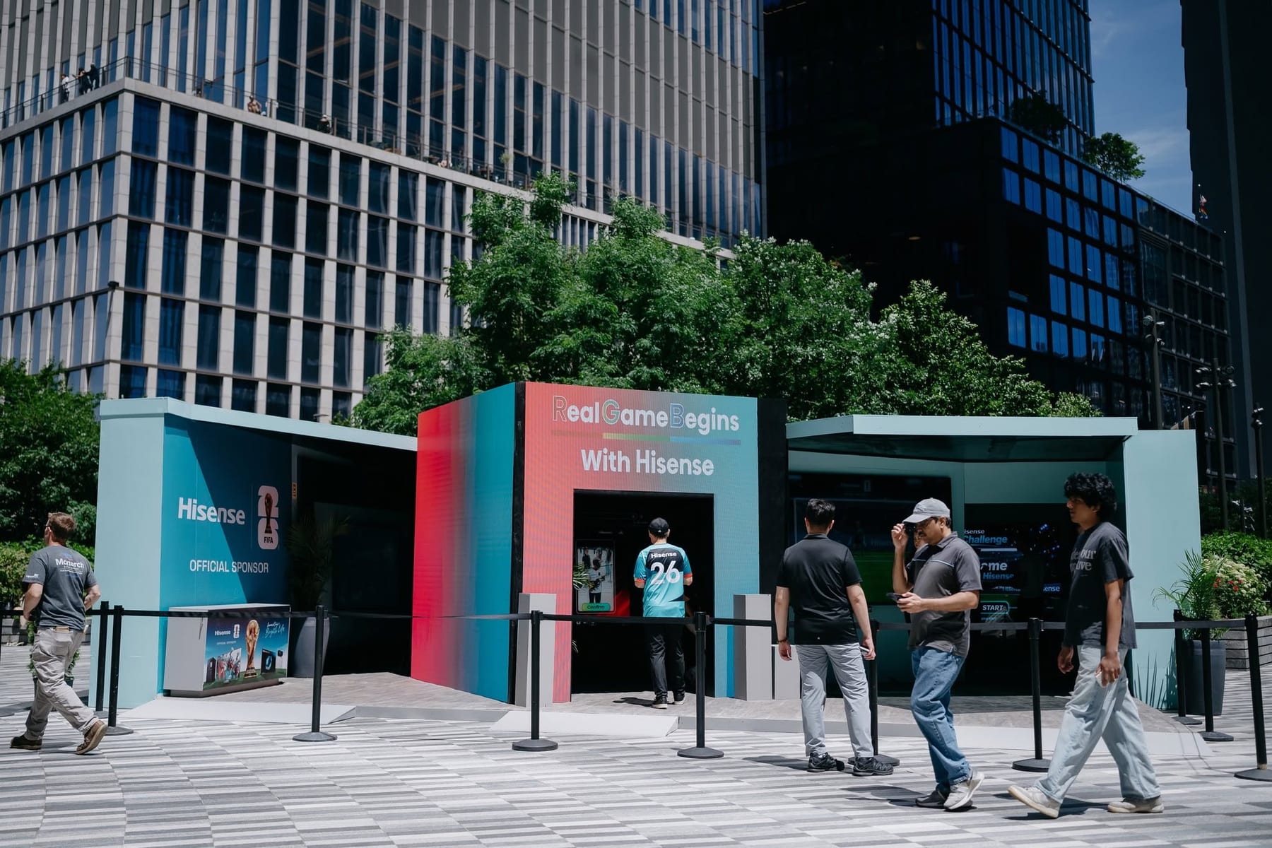 The pop-up pavilion seen straight on, its three bays carrying the Real Game Begins With Hisense wordmark, with pedestrians crossing the Hudson Yards plaza in front